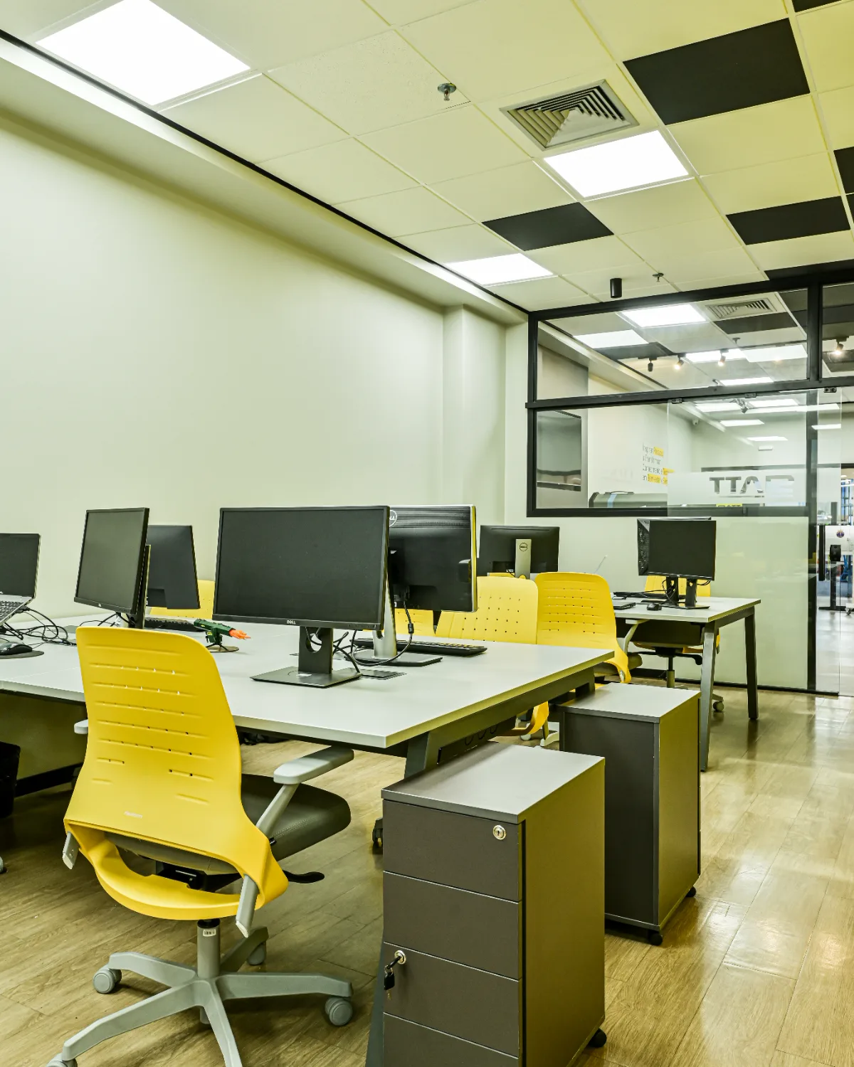 Conjunto de estações de trabalho com gaveteiros móveis, forro em xadrez e sala envidraçada ao fundo