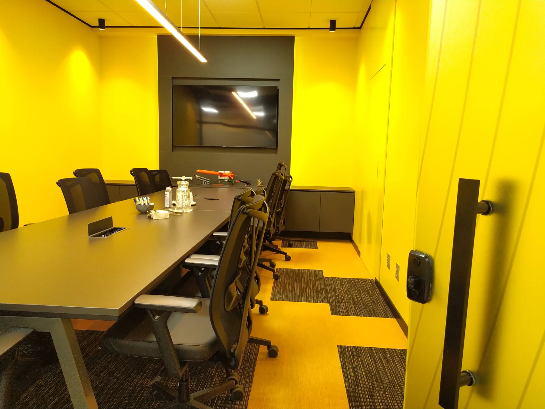 Sala de reunião executada em amarelo integral, com mesa longa, monitor sobre painel escuro e piso em placas mescladas