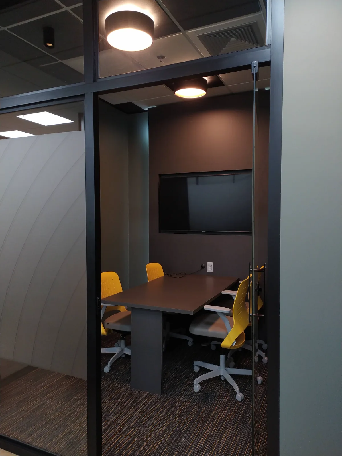 Sala de reunião menor, fechada em vidro, com mesa para quatro lugares, monitor e iluminação de teto pontual