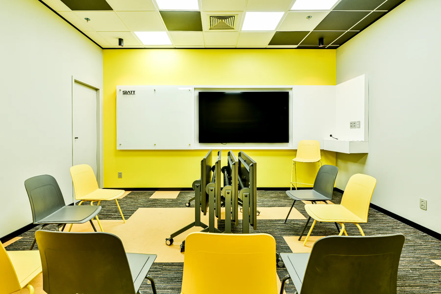 Espaço para workshops executado, com mesas dobráveis recolhidas, cadeiras empilháveis, quadro branco e monitor na parede amarela