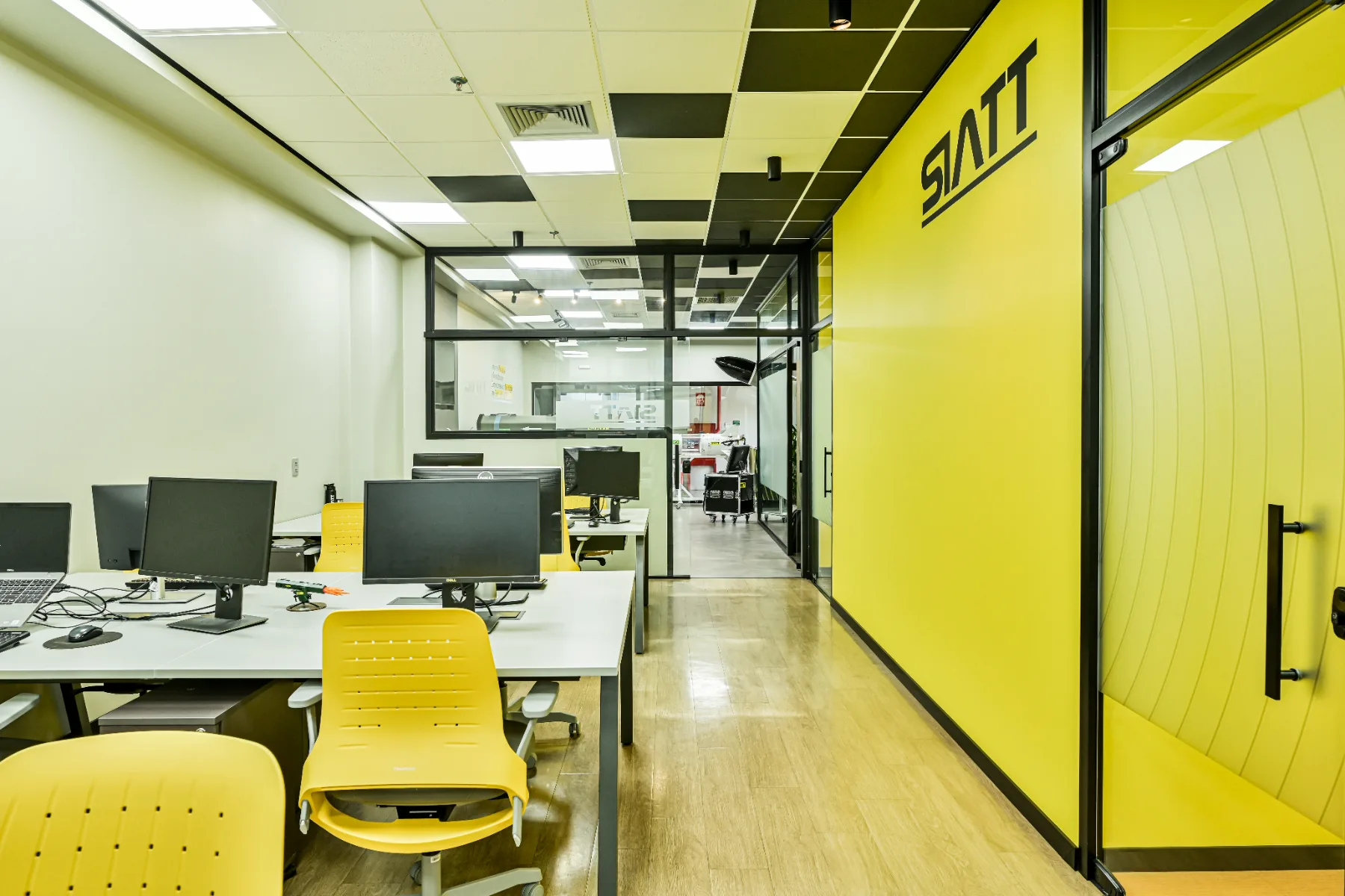 Área de desenvolvimento e operação do SIATT Trade, com estações de trabalho, vedações em vidro e parede amarela com a marca aplicada