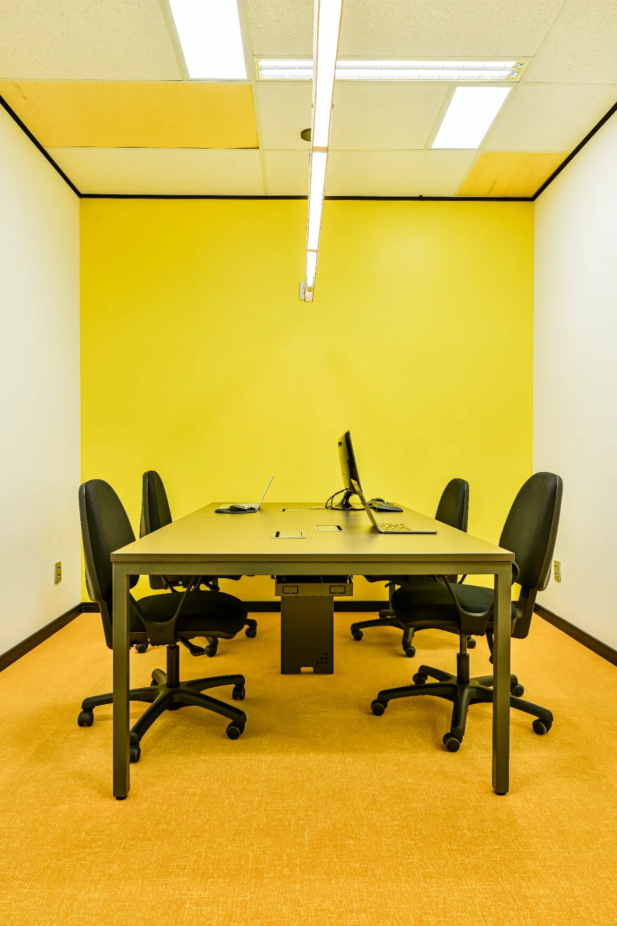 Sala de meeting com parede amarela, mesa para quatro pessoas e piso em carpete