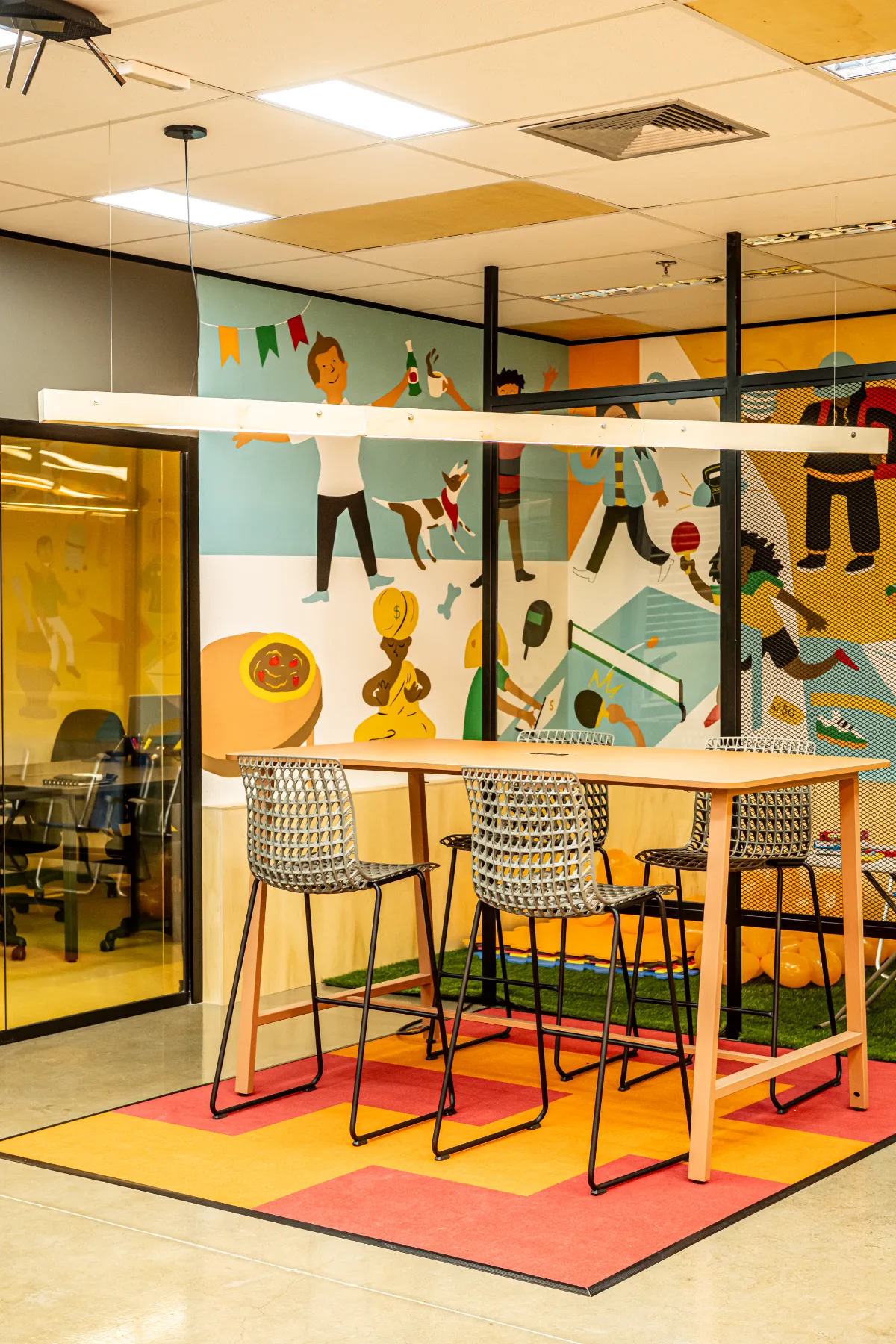 Mesa alta com banquetas diante do mural ilustrado, ao lado da sala de reunião envidraçada