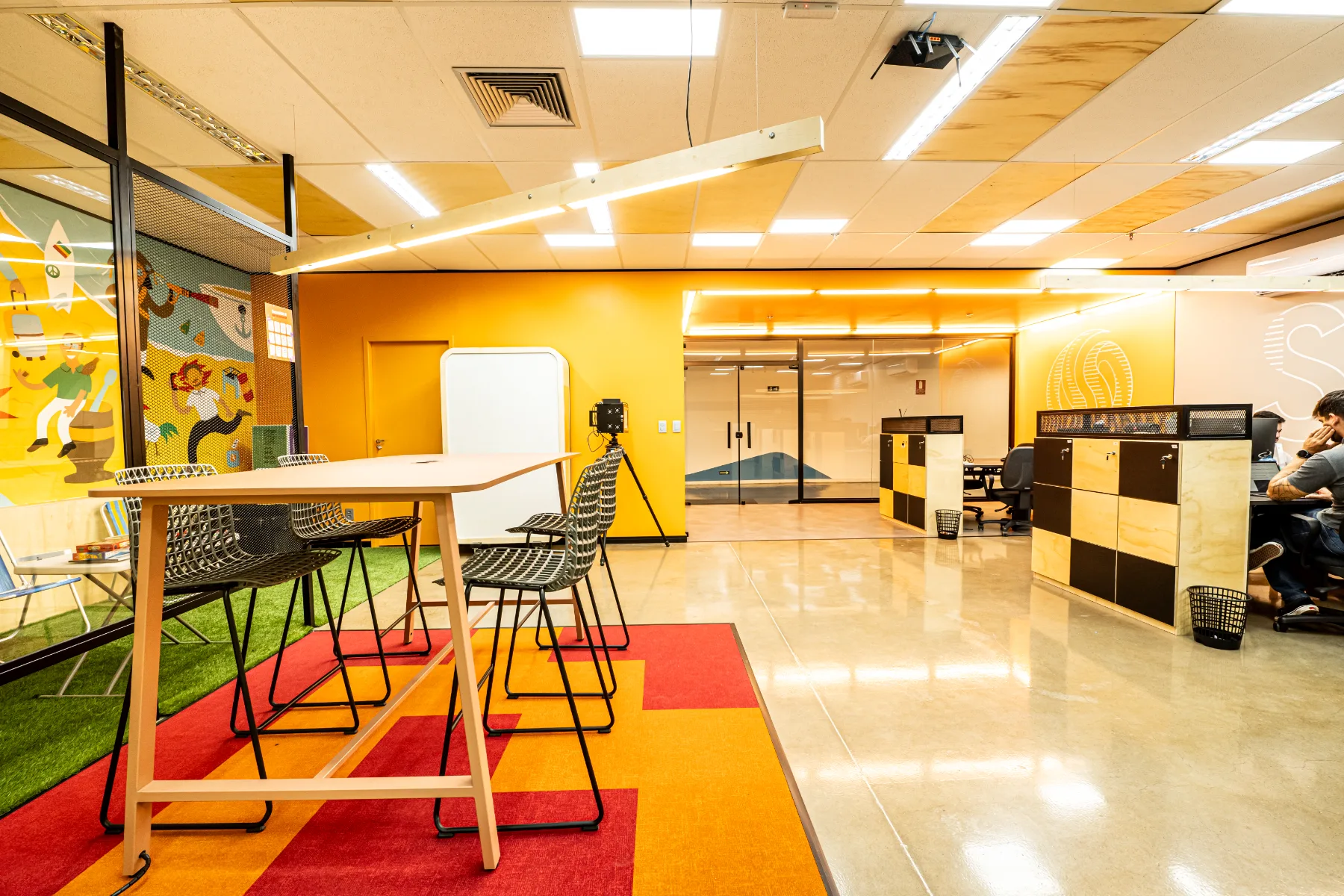 Área de descompressão com mesa alta sobre tapete colorido, grama sintética e, ao fundo, o open space com armários de compensado