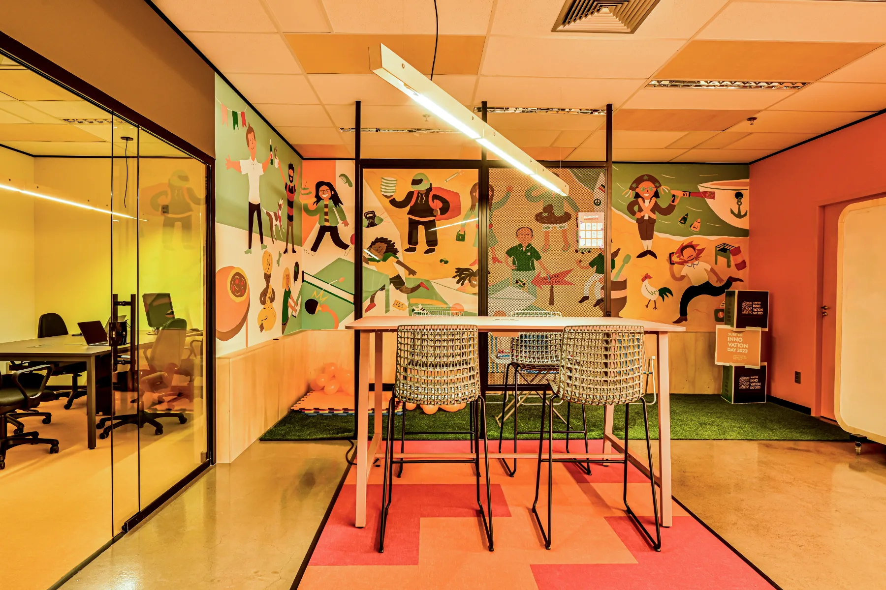 Área de convivência com murais ilustrados, mesa alta com banquetas e sala de vidro âmbar ao lado